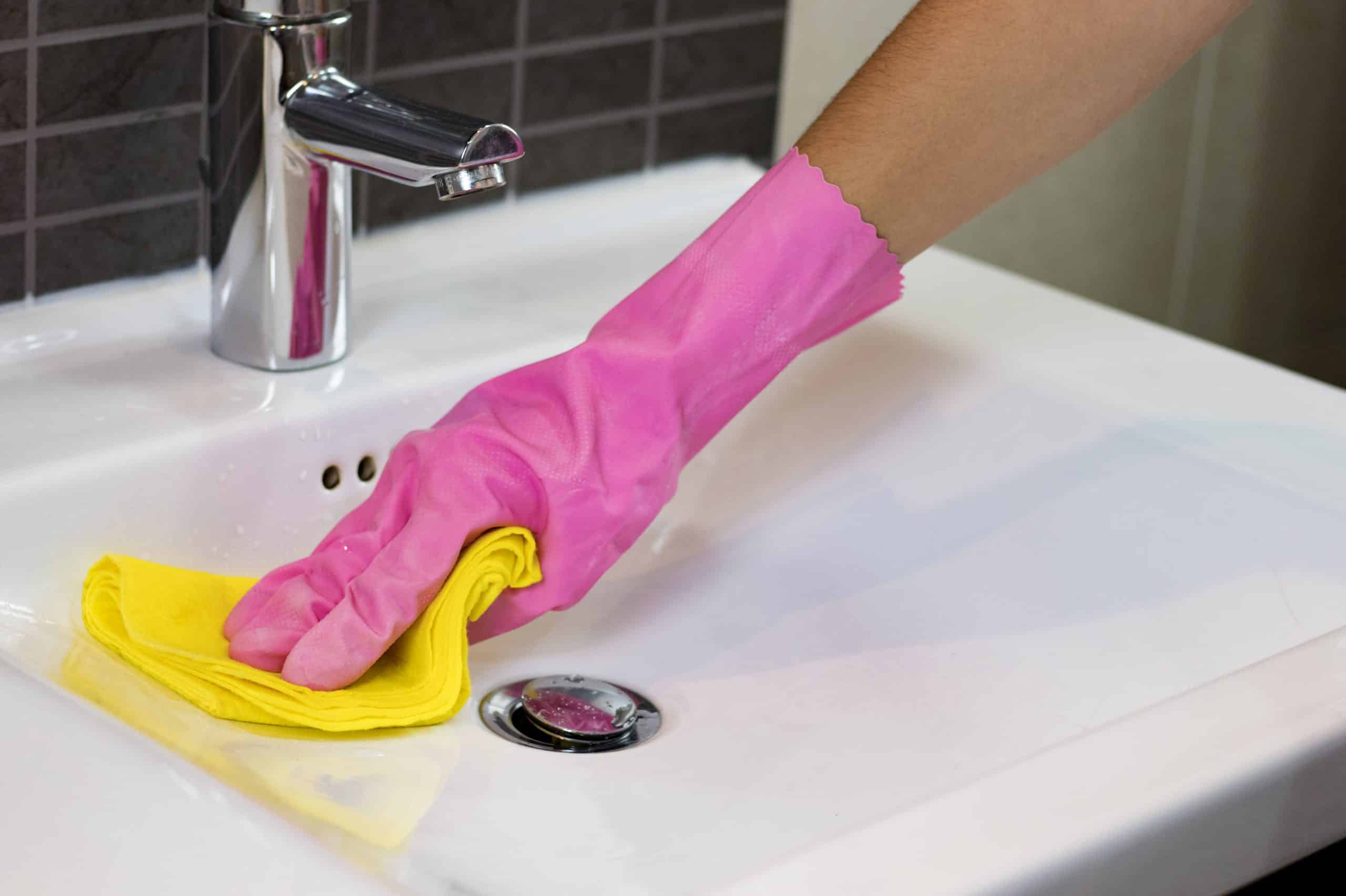 Closeup of a person wearing pink rubber gloves cleaning a bathroom sink with a cloth