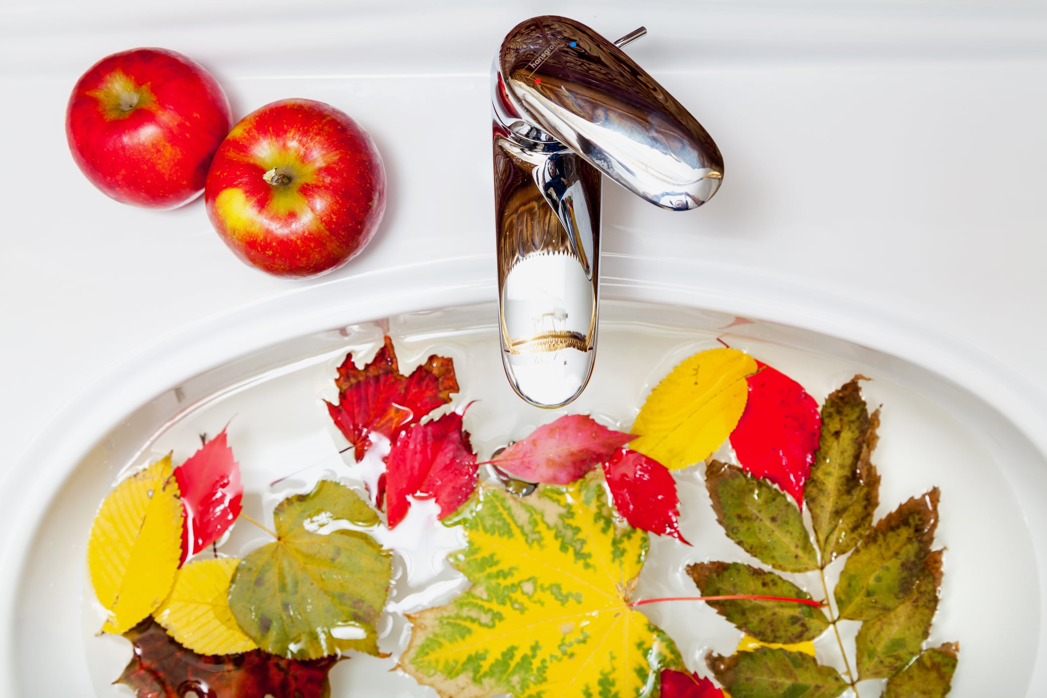 A white porcelain bathroom sink with fall foliage floating inside it and two red apples next to the faucet.