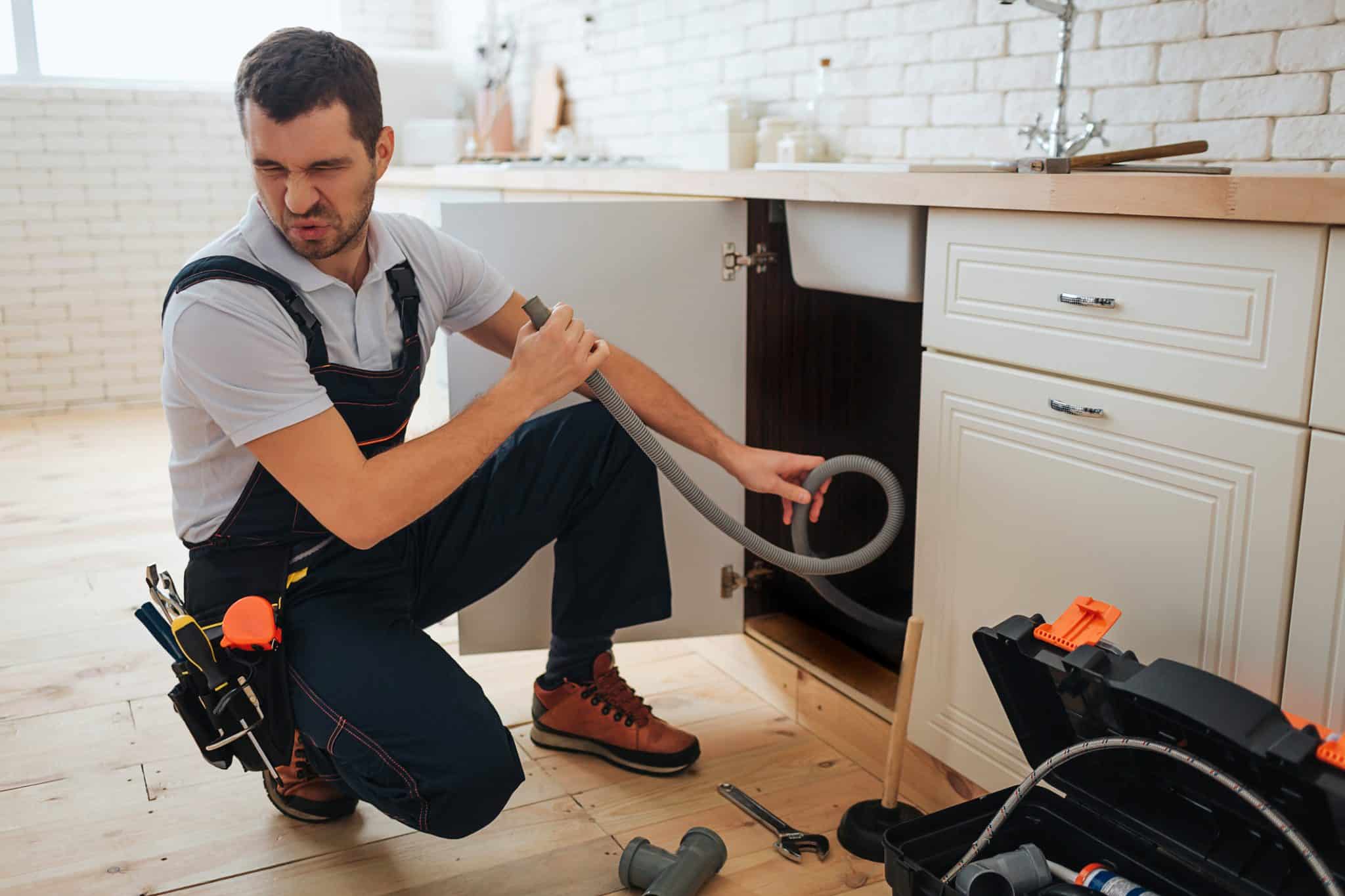 Young plumber recoils from rotten egg smell water while inspecting pipes for sewer smell in San Francisco house.